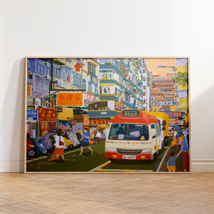 A large framed canvas print of a vibrant, colorful street scene with a red and white bus, leaning against a white wall on a light wood herringbone floor.