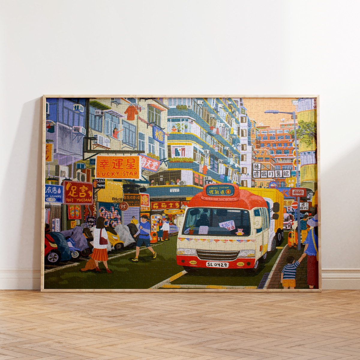 A large framed canvas print of a vibrant, colorful street scene with a red and white bus, leaning against a white wall on a light wood herringbone floor.