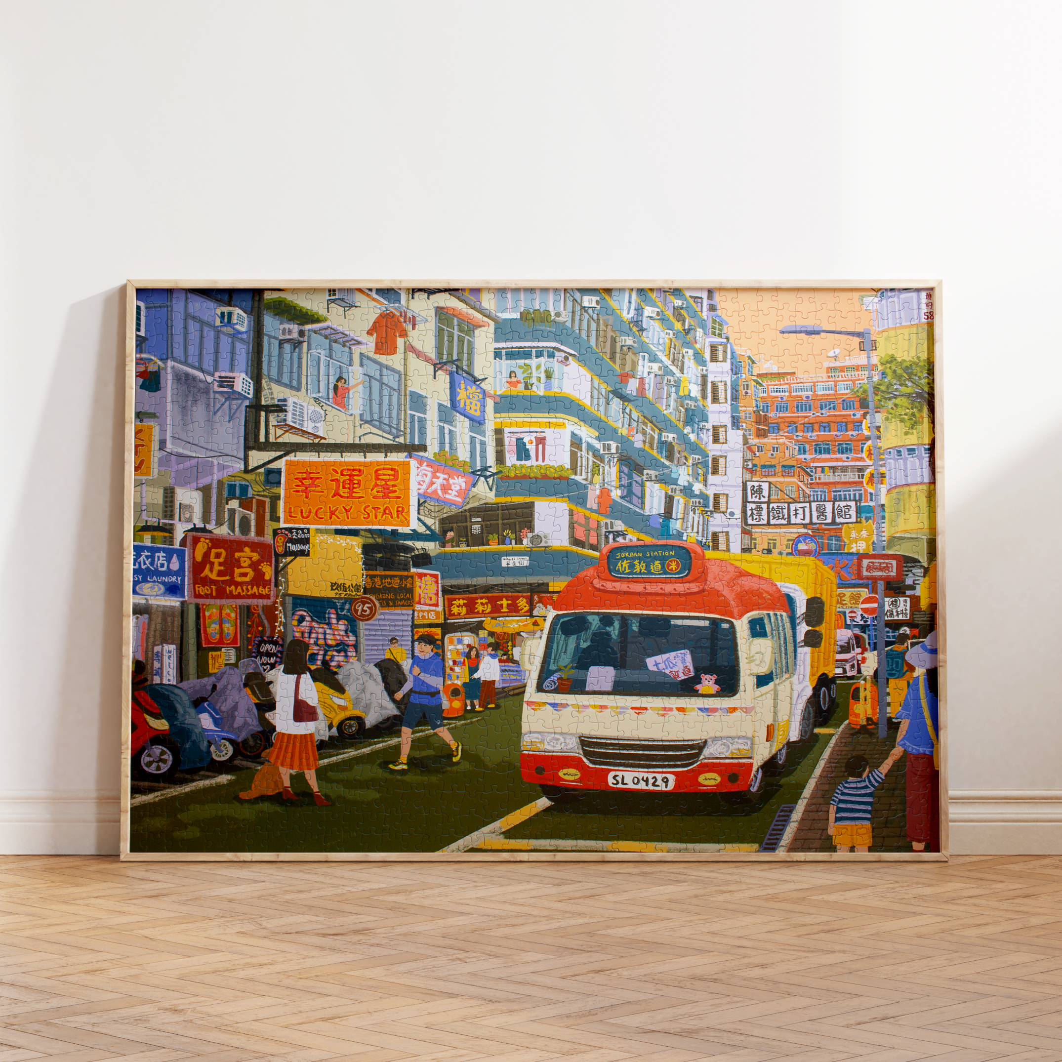 A large framed canvas print of a vibrant, colorful street scene with a red and white bus, leaning against a white wall on a light wood herringbone floor.