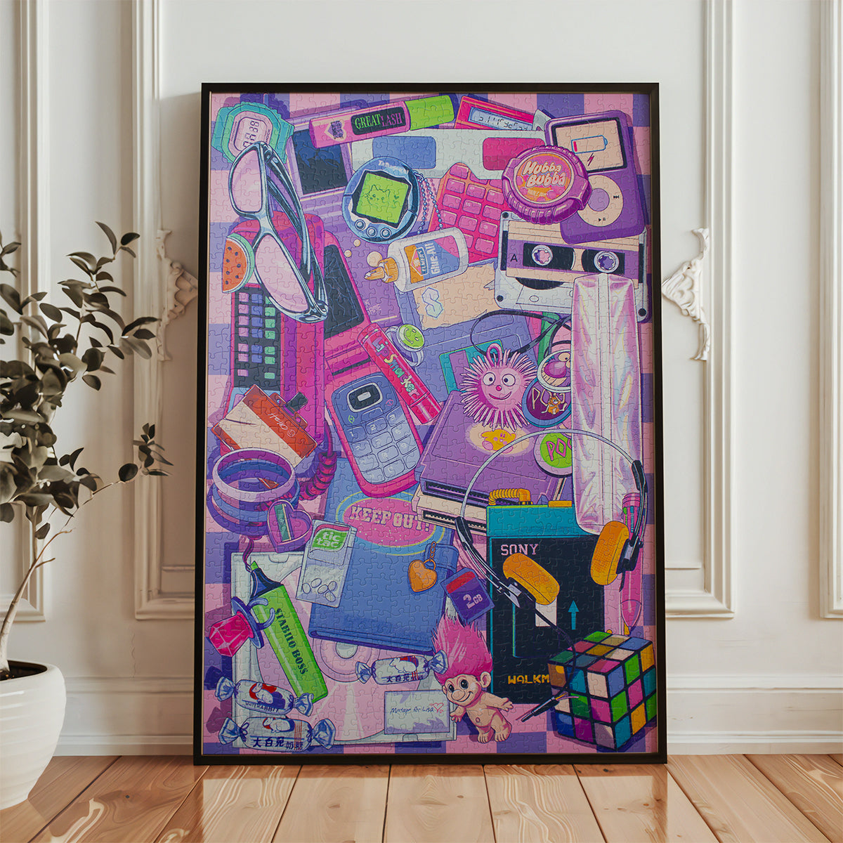 A framed art print featuring a colorful collage of pop culture icons, retro gadgets, and playful illustrations in pink, purple, blue, and yellow tones, leaning against a white paneled wall on a light wood floor next to a potted plant.