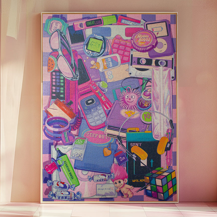 A large framed art print leaning against a pink wall in a softly lit room. The artwork is a dense collage of colorful everyday objects and icons, including a pink phone, cassette tape, calculator, sunglasses, headphones, and other small illustrated elements.